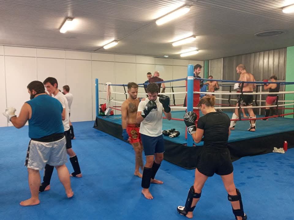 La salle du club pendant un cours adultes : des pratiquants travaillent par deux autour du ring, gants aux poings, d'autres s'échauffent sur les tapis.