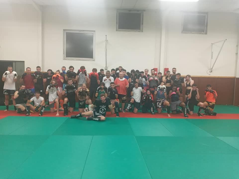 Photographie de groupe dans le gymnase : une centaine d'adhérents, enfants et adultes, rassemblés sur les tapis à la fin d'un entraînement.